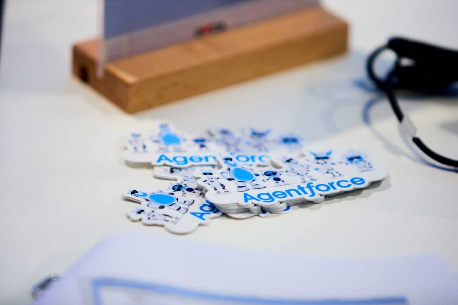 Stickers met het woord "Agentforce" in blauwe letters zijn op een wit oppervlak geplakt, naast een zwarte kabel en een houten standaard.