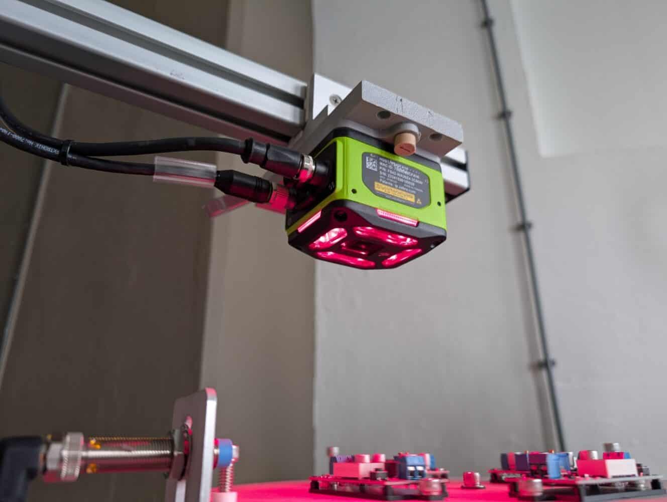 Zebra NS42 Smart Vision Sensor Close-up van een groene industriële camera met rode lampjes, gemonteerd op een metalen frame, gepositioneerd boven elektronische printplaten.